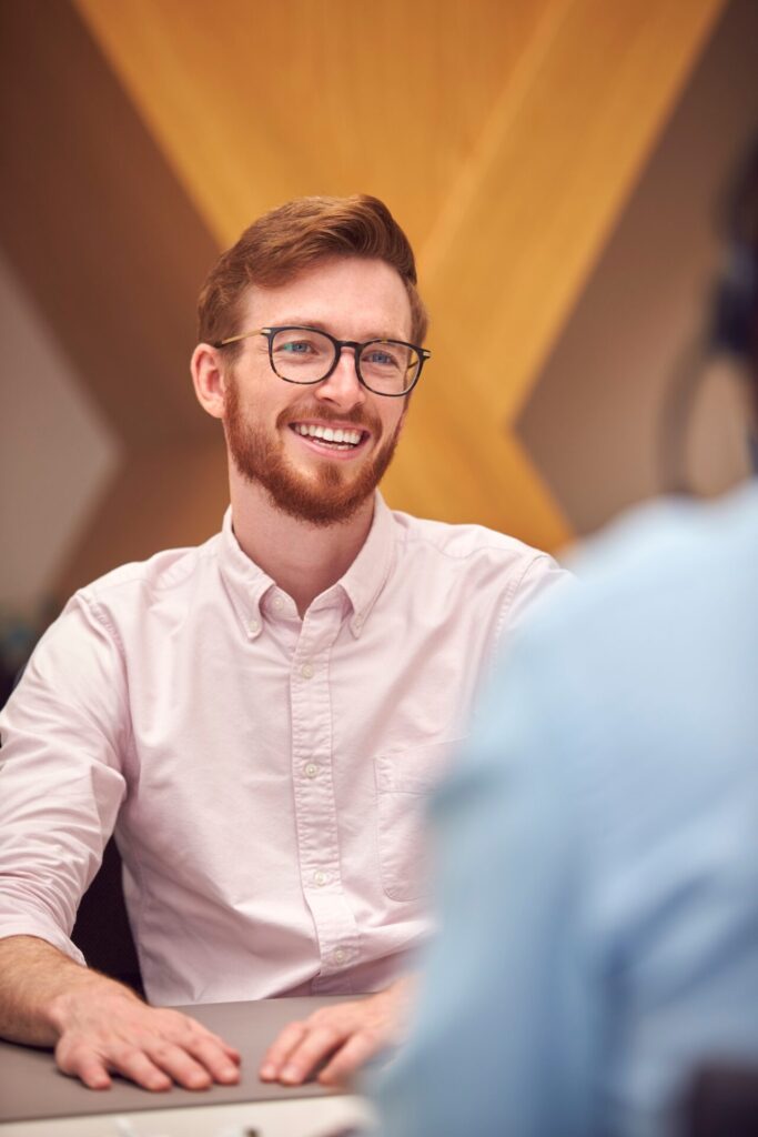 Businessman In Office Talking To Male Colleague Sitting At Desk